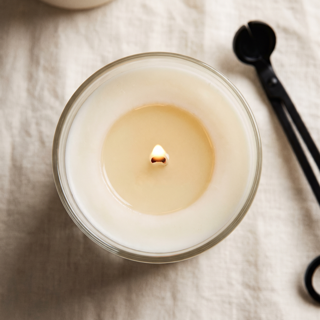 A candle seen from directly above with a full melt pool reaching the edge of the glass and a wick trimmer resting beside it