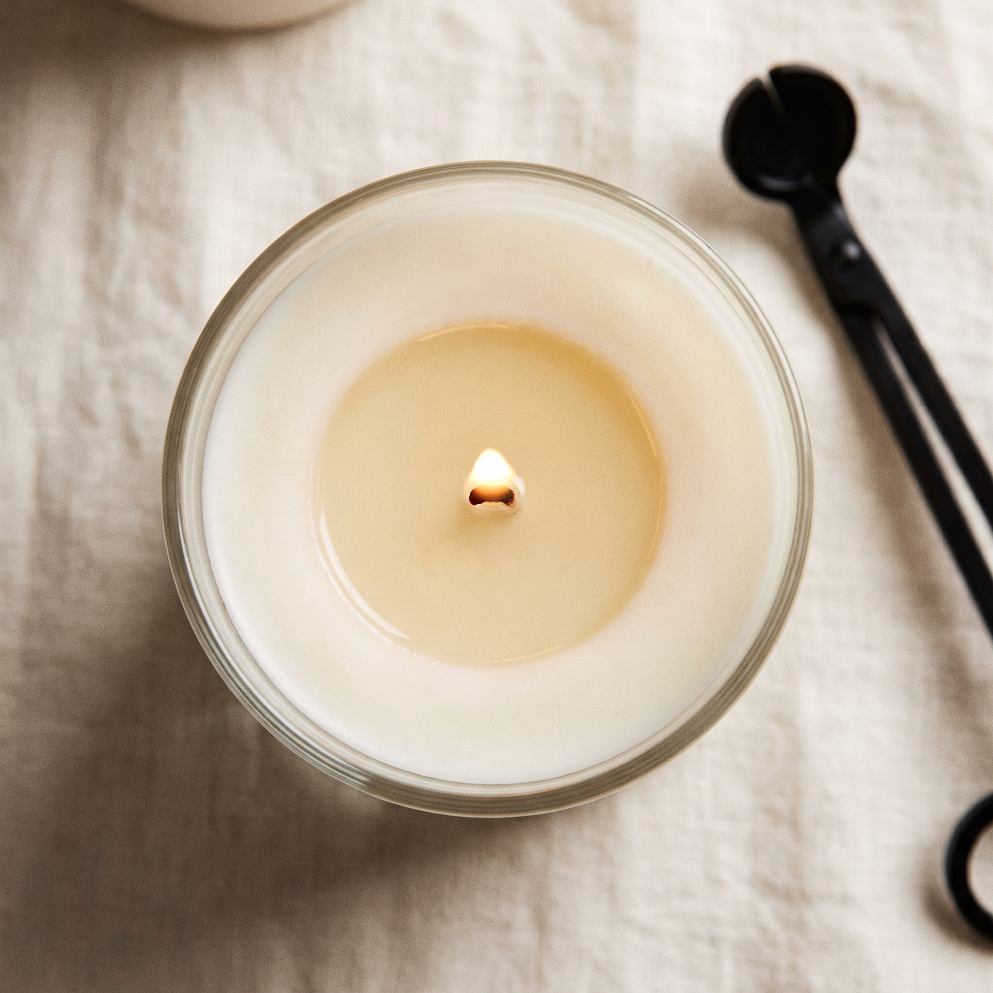 A candle seen from directly above with a full melt pool reaching the edge of the glass and a wick trimmer resting beside it