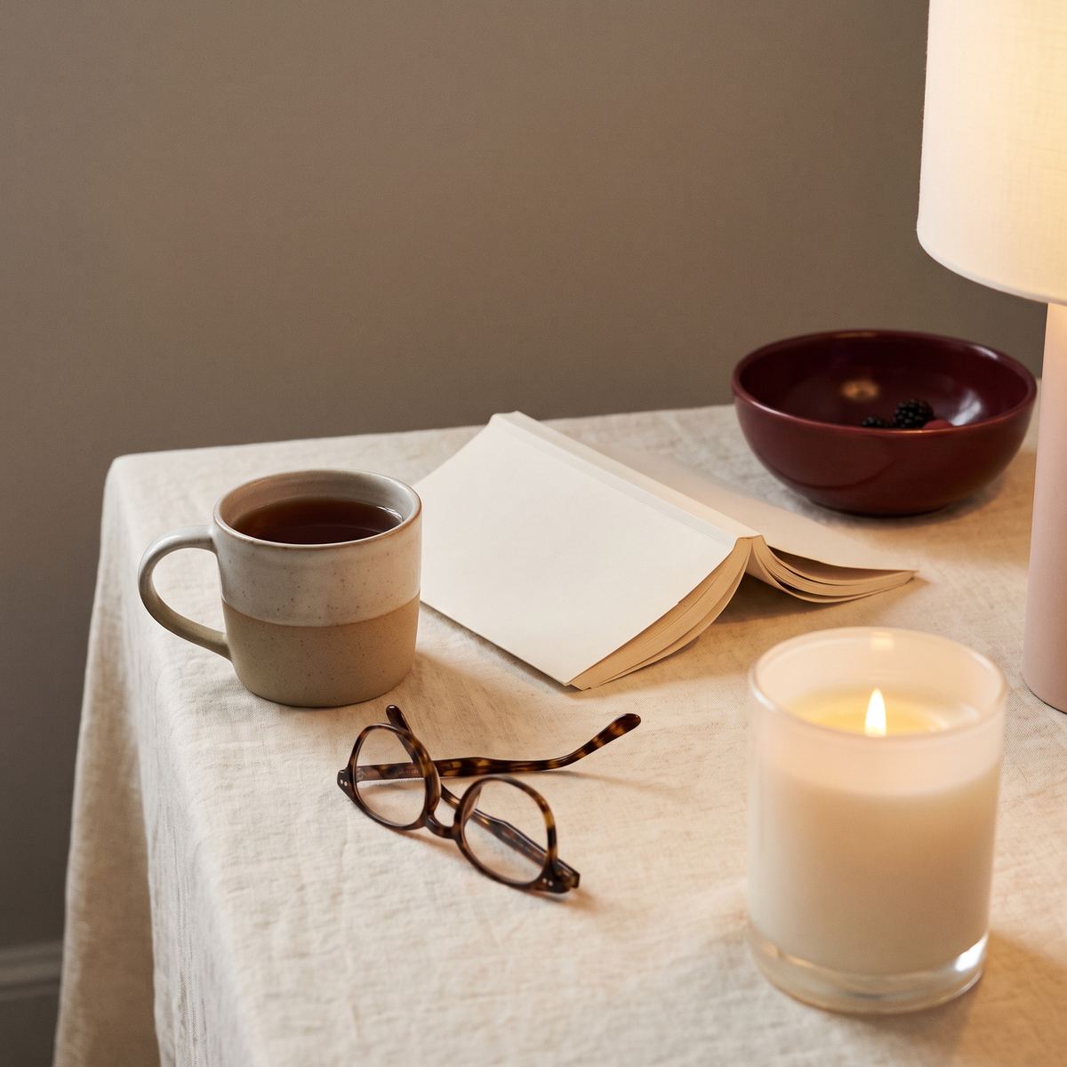 A half-drunk mug of tea, a book lying open face down and reading glasses on linen, with a candle lit in the foreground